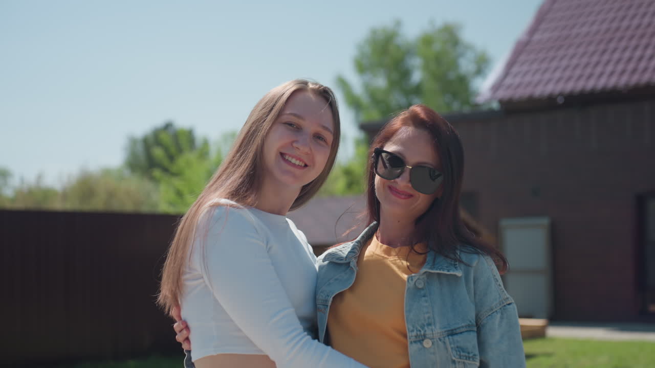 Two joyful women reconnect and embrace with genuine smiles outdoors on bright sunny day, standing on green lawn in front of modern house with red tiled roof and wooden cabin structure behind