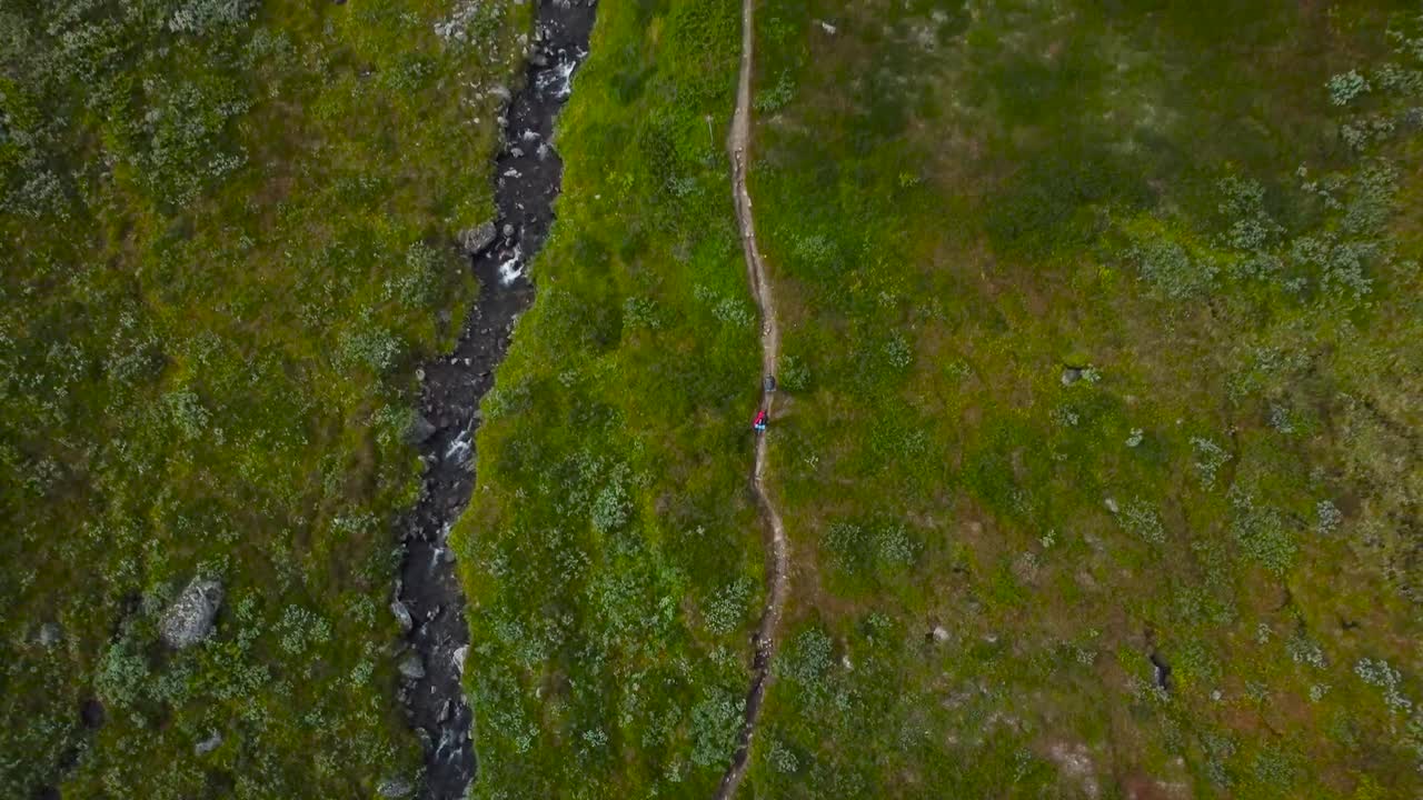 Top down aerial view perspective of two hikers or backpackers walking on a rural muddy road in Sweden green grassy and brown fields landscape while a narrow river flows besides them with white water.