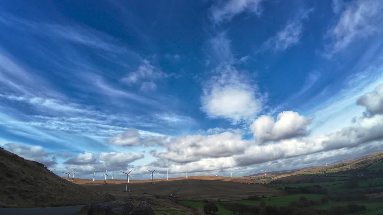 Ultra Wide View of Welsh Wind Farm Over Moorland with Dozens of Farmed Green Fields with Contrasting Puffy Clouds and Blue Sky