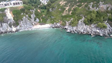 Luftaufnahme-Des-Kleinen-Türkisfarbenen-Strandes-Mit-Felsen,-Unter-Einem-Gebäude-Mit-Heißen-Quellen,-In-Der-Gegend-Von-Agia-Paraskevi,-Chalkidiki,-Seitwärtsbewegung-Per-Drohne