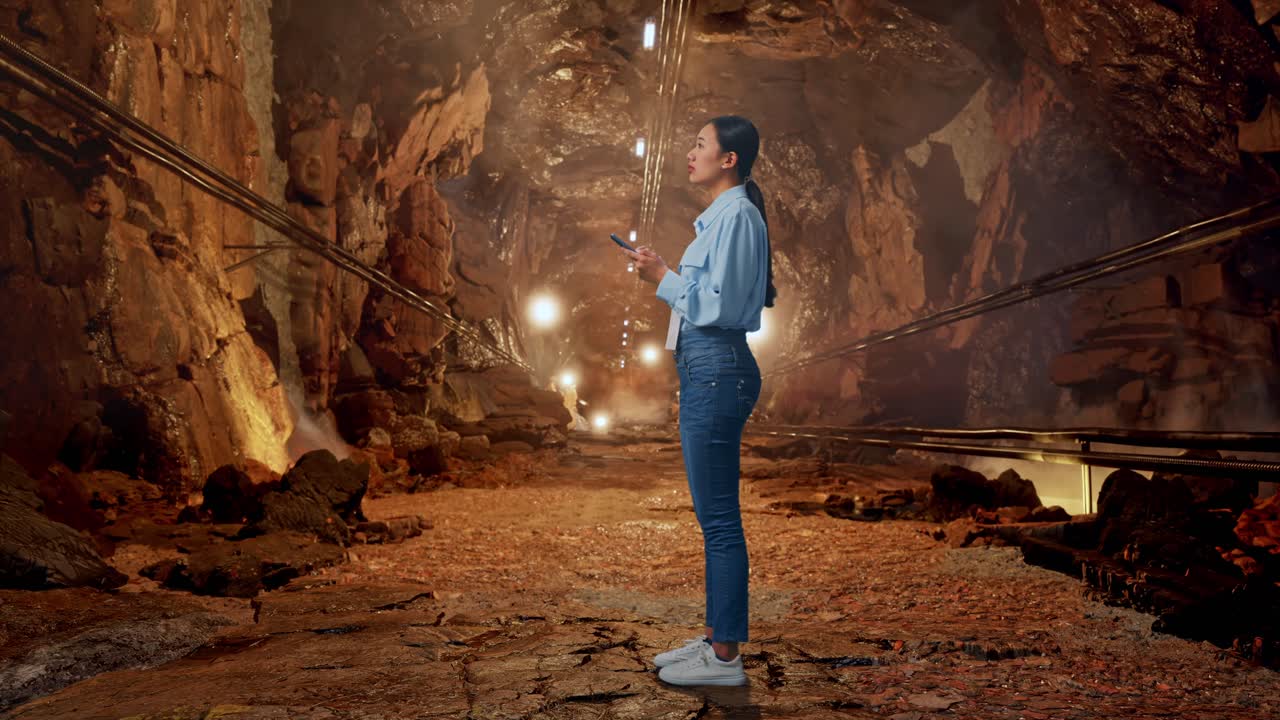 Full Body Side View Of Asian Female Worker With Her Phone In Underground Mine Tunnel, She Observes By Looking Around Before She Come To Concentrating With Her Smartphone