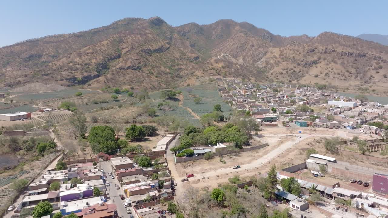 Blue agave agricultural fields in Amatitan town, ingredient for tequila production, residential houses and rugged mountain landscape in view, establishing drone shot