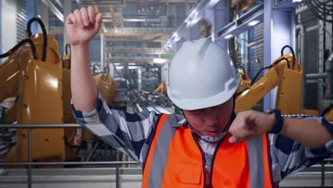 close up of asian male engineer with safety helmet screaming goal and dancing celebrating with industrial robotic arms working