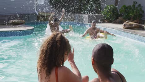 Two-diverse-female-friends-talking-in-swimming-pool-while-their-male-friends-jumping-into-water