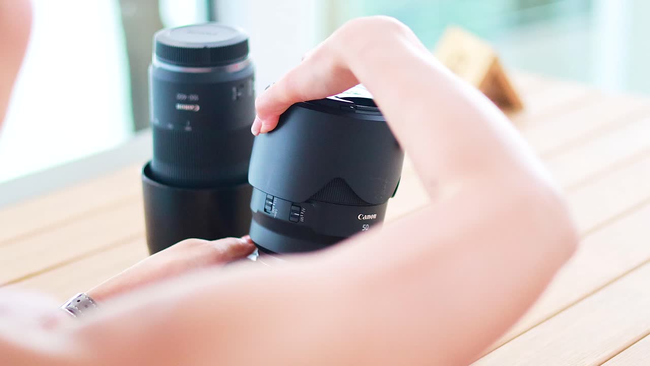 A woman changes a camera lens on a wooden table in bright, natural light, highlighting photography equipment handling