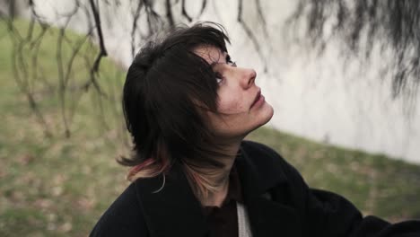 young woman in a black coat standing under a windy tree in a park in autumn in afternoon-5