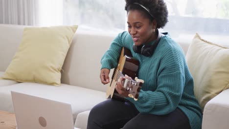 Feliz-Mujer-Afroamericana-Practicando-Tocar-La-Guitarra-Usando-Una-Computadora-Portátil-En-Casa,-Cámara-Lenta