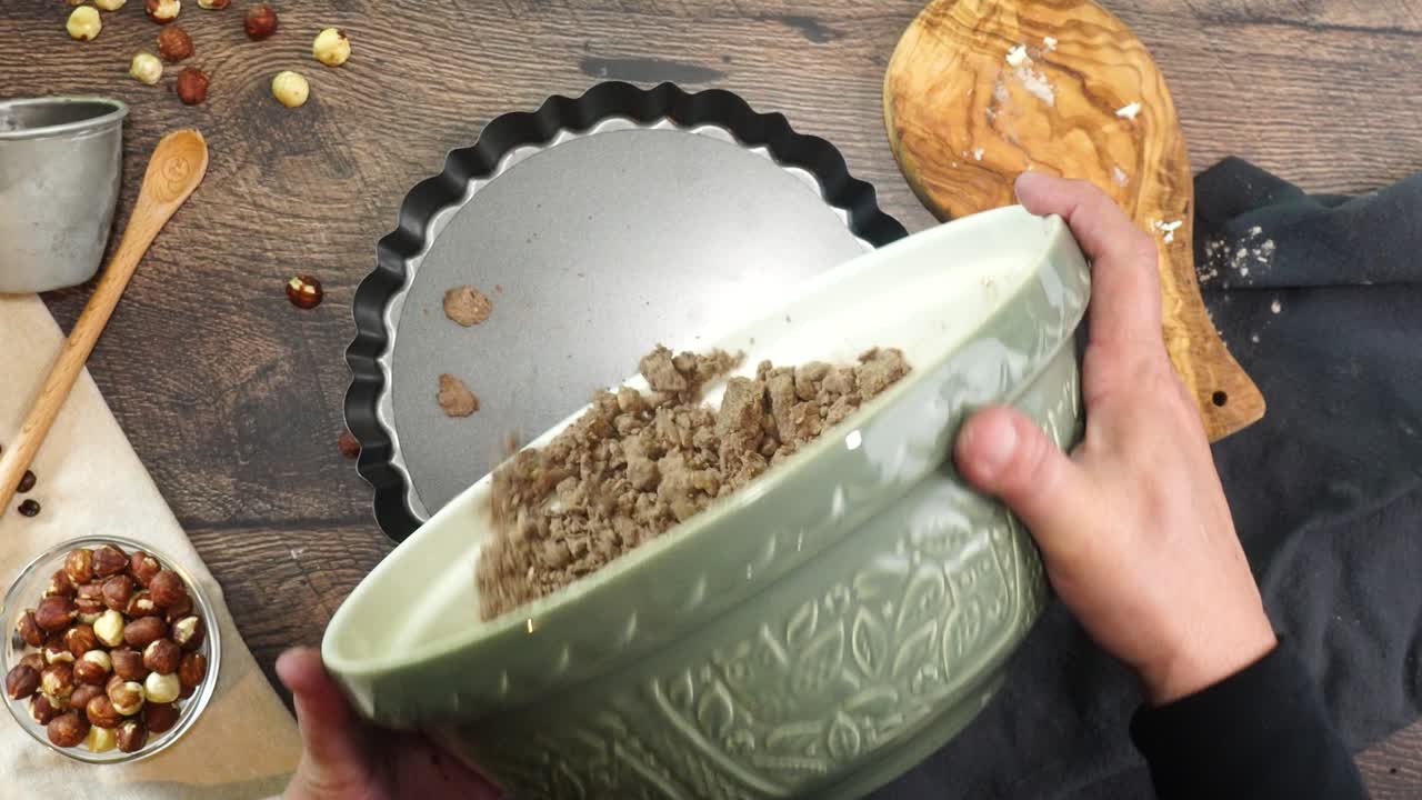A delicious dessert, Coffee Hazelnut Cream Tart. Made with espresso powder, chocolate crust and loaded with crushed hazelnut. Hand mixing chocolate pie crust in a clear mixing bowl on a wooden table