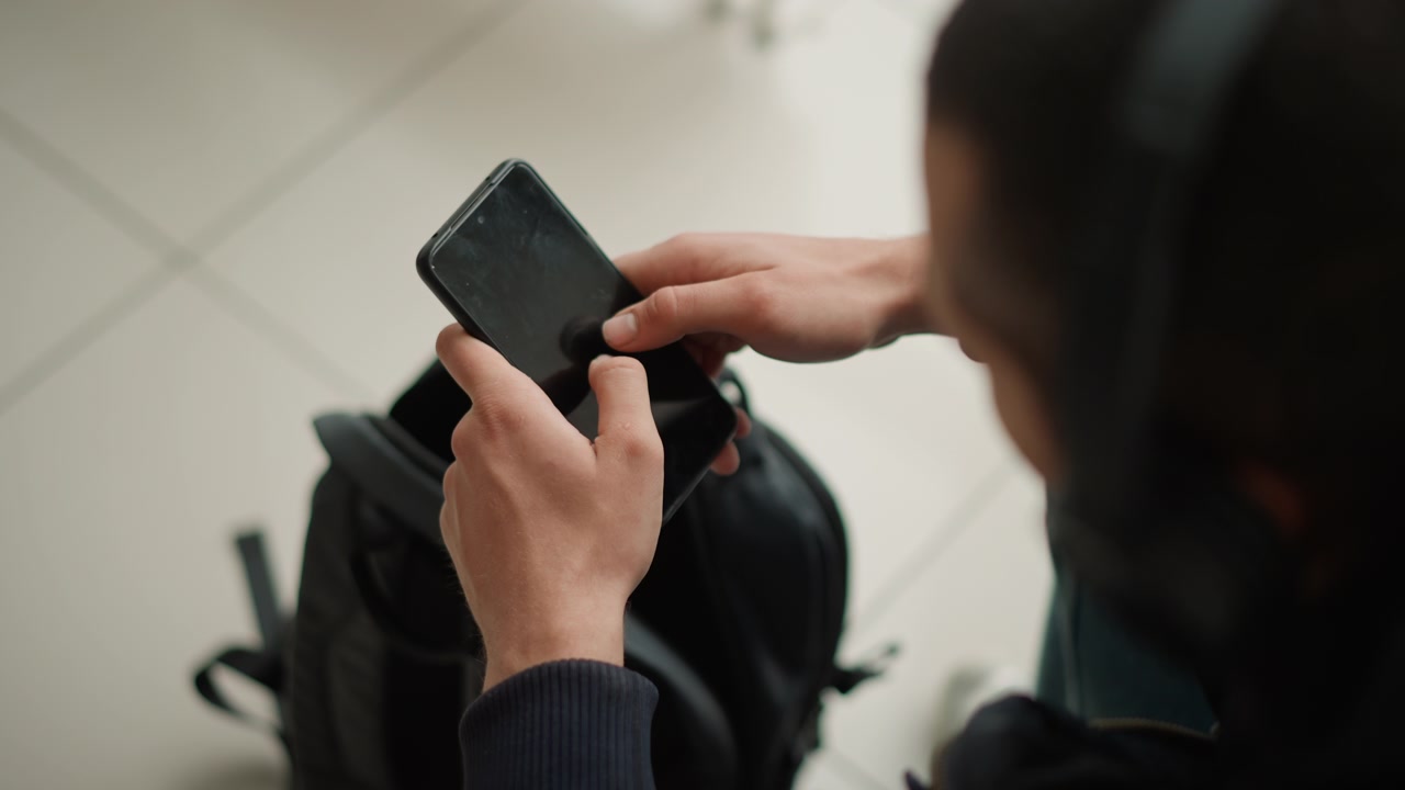 Close up of person holding and interacting with smartphone focusing on hands tapping screen. black backpack is visible in background on tiled floor. Soft lighting creates natural indoor
