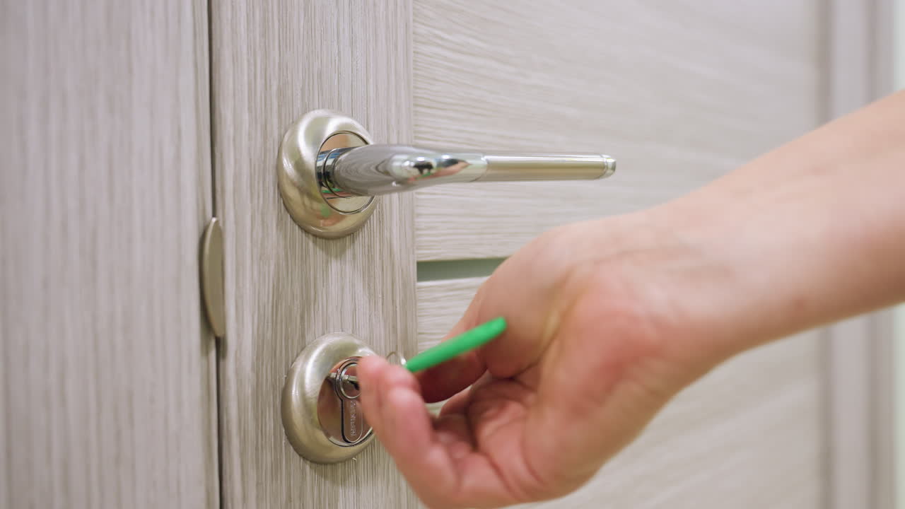 Close up of female hand inserting key into modern metallic door lock preparing to unlock light wood-textured door with chrome handle indoors showing concept of safety access privacy security control