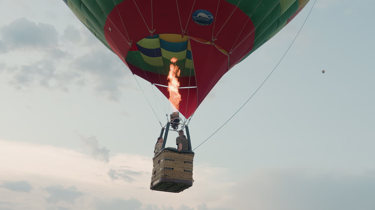 couple riding wicker basket gripping burner controls while vibrant flame roars overhead under pastel sunset sky distant balloon speck on horizon capturing dynamic aerial