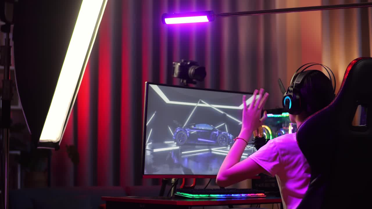 vista trasera de una chica asiática que juega a un juego de carreras de coches a través de la red en la computadora, hablando con los participantes en el micrófono. videojuego de transmisión en vivo, escritorio iluminado por luz de tira led rgb
