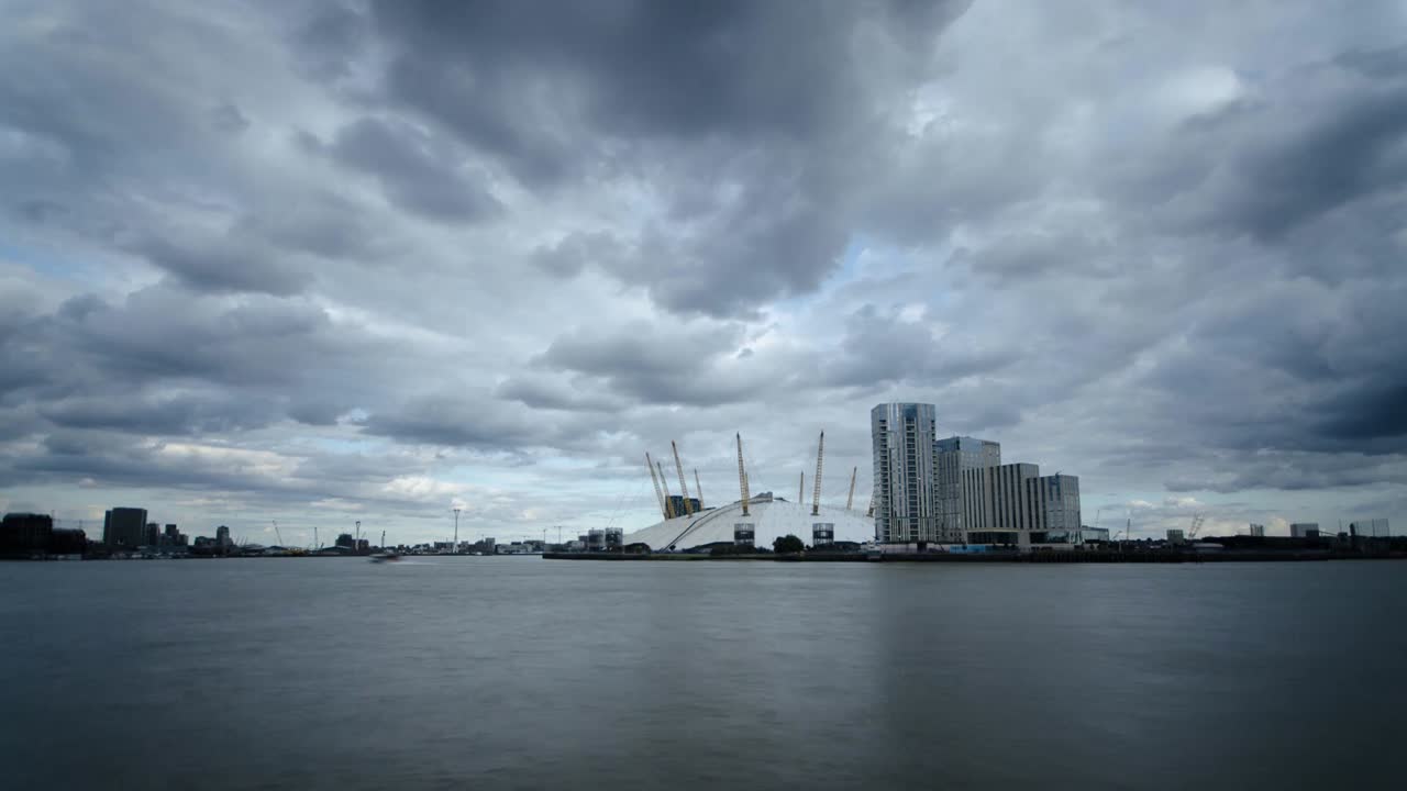 Timelapse captures the iconic O2 Arena along the River Thames, set against dramatic, shifting clouds. The wide-angle view emphasizes the imposing architecture and moody atmosphere