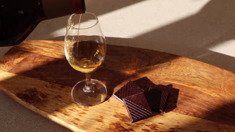 pouring single malt scotch whisky into a copita glass, paired with squares of dark chocolate on a wooden serving board, with shadows from the evening sun