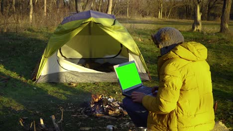 un joven trabaja en una computadora portátil cerca del fuego en el bosque