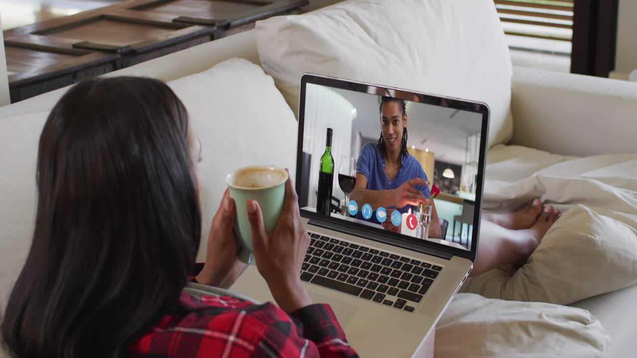 African american woman holding a coffee cup having a video call on laptop sitting on couch at home
