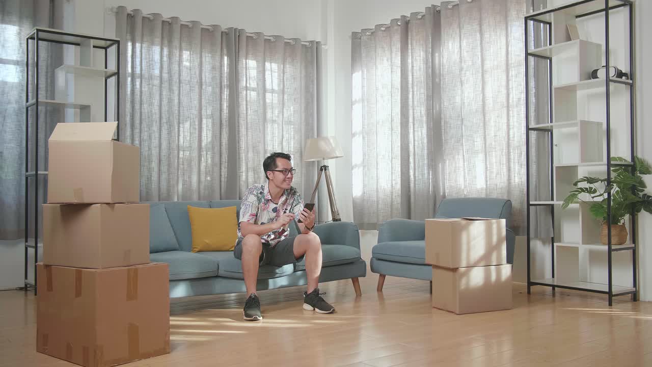 소파에 앉아 새 집으로 이사한 후 스마트폰에 있는 사진과 집을 비교하는 카드보드 상자를 가진 아시아인
