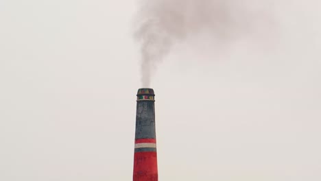 red and black brick field chimney emitting dark smoke with a grey sky in the background, bangladesh