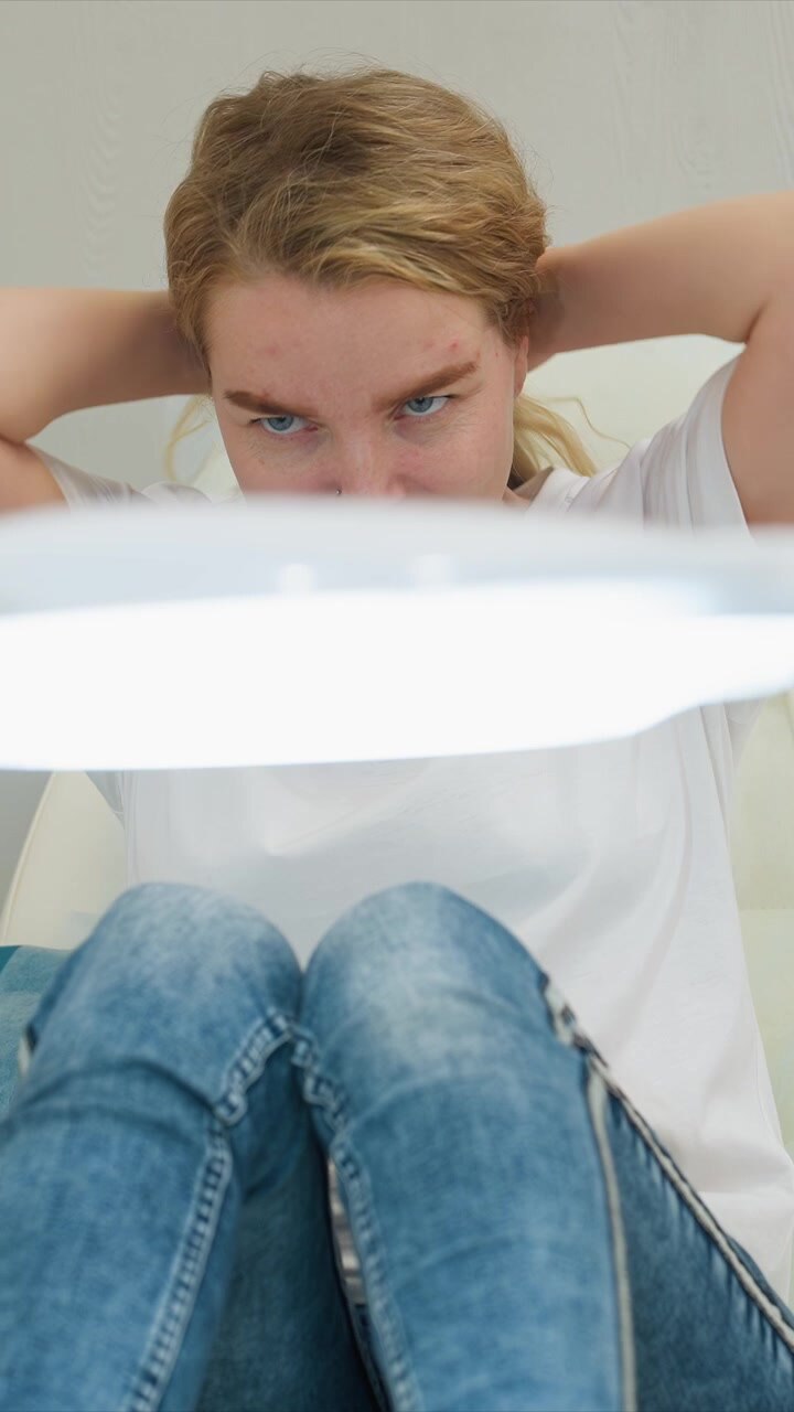 Close up shows lower body details blue jeans with visible stitching white fabric texture of chair. woman hands rest on her lap under bright light suggesting woman is preparing