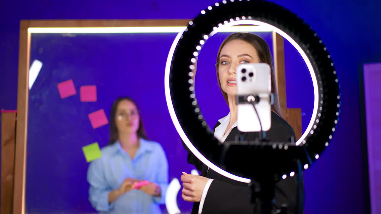 Studio brainstorming session. A woman near a ring light observes as another writes on sticky notes at a glass board during brainstorming