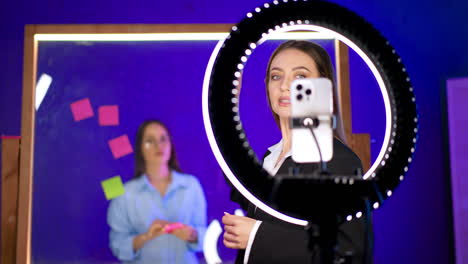studio brainstorming session. a woman near a ring light observes as another writes on sticky notes at a glass board during brainstorming