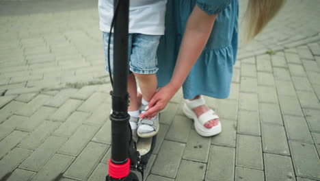 primer plano de la mano de una mujer guiando el pie de un niño a la plataforma del scooter mientras levanta su pierna izquierda