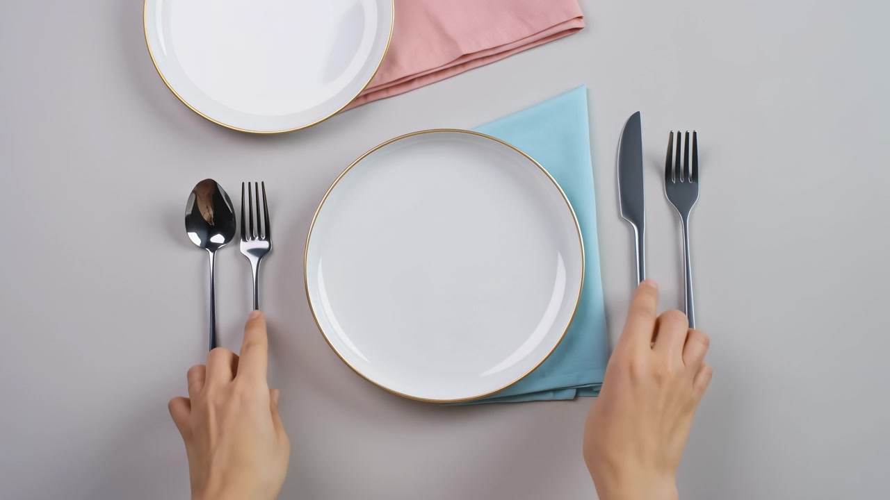 Top view of modern dining table setup with hands adjusting central plate beside polished spoon, fork, and knife, folded pastel napkins, smooth ceramic plates, and calm hospitality styling under soft