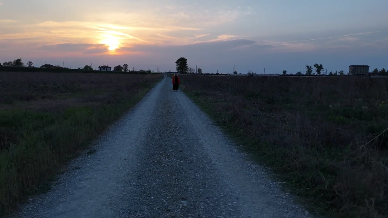 夕暮れ時、広々とした農地を抜ける田舎の砂利道を一人で歩く人物。孤独と瞑想を表現し、ドローンが後退する