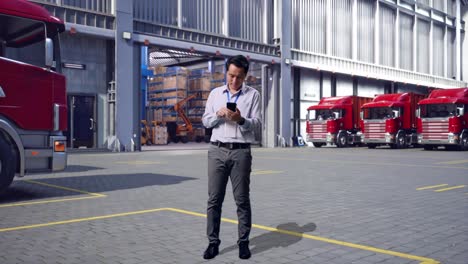 full body of an asian male professional worker standing with his smartphone, outside of logistics distributions warehouse, working continuously