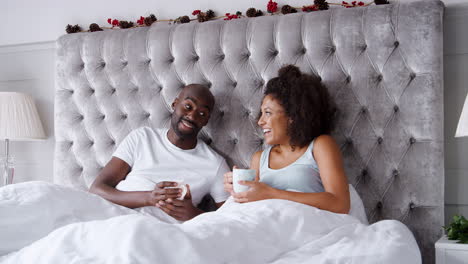 Young-adult-black-couple-sitting-up-in-bed-relaxing-with-cups-of-coffee,-close-up