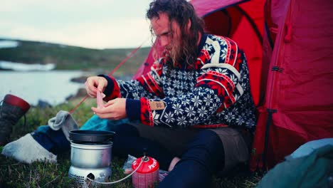 a man cooking on a portable stove while camping by the lakeshore along the route from skurven to mefjellsvatnet in indre fosen, trøndelag, norway - close up