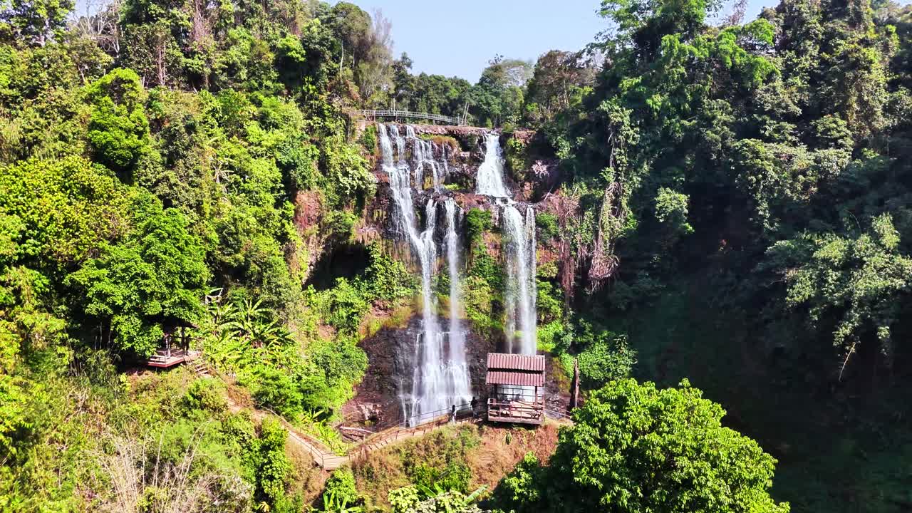 Tad Gneuang Waterfall in southern Laos plunges over rocky cliffs into dense tropical forest, with wooden viewing huts and a winding trail inviting peaceful exploration of the natural surroundings