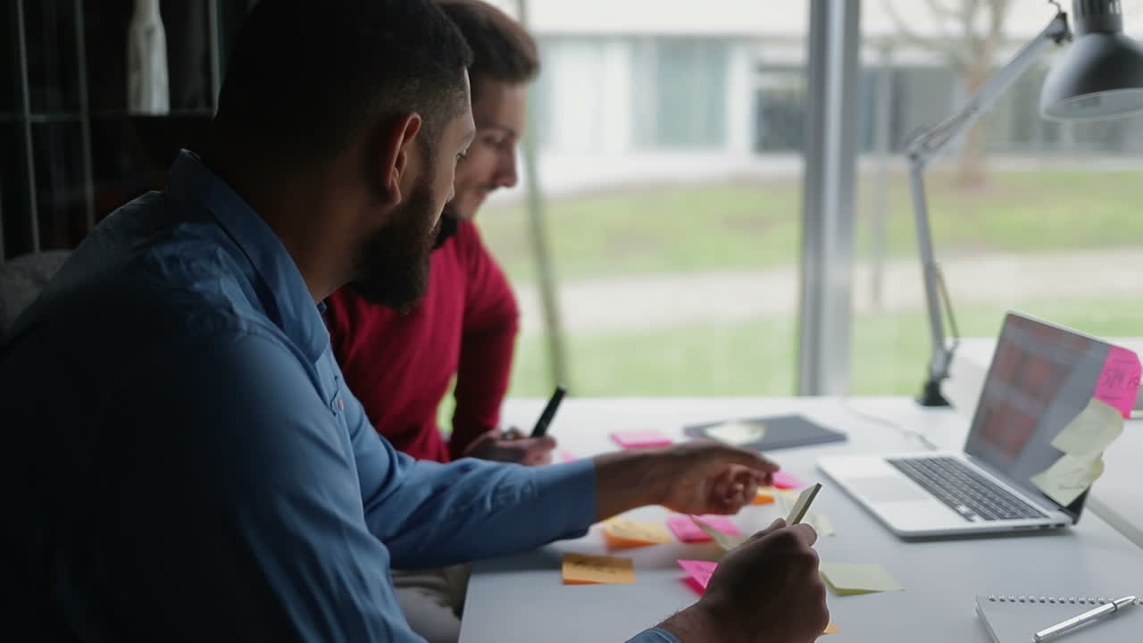 dos hombres jóvenes sentados en una mesa con notas adhesivas y una computadora portátil.