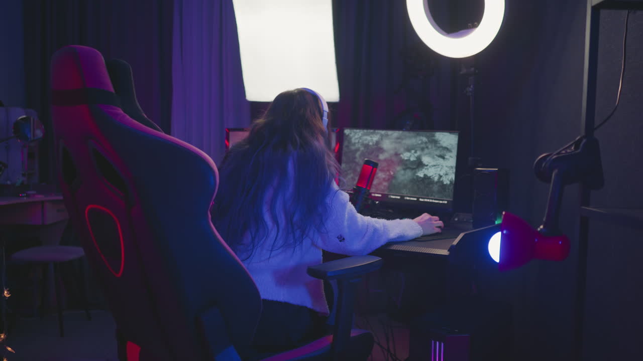 rear view of woman with long hair sitting on gaming chair playing video game on desktop monitor with headset on, surrounded by lighting gear, microphone, and other equipment in purple-lit studio