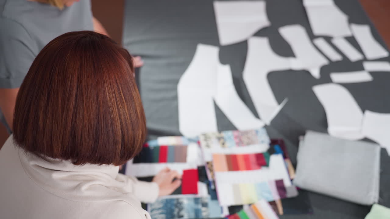 Aerial view of two weavers touching fabric samples and color swatches, analyzing textile options, bright workspace filled with cut pattern pieces, creative preparation for fashion