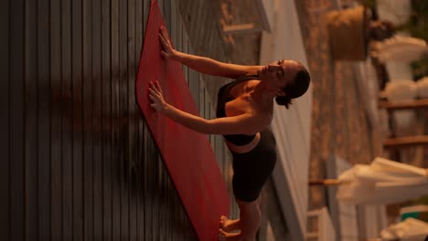 Vertical-video:-A-brunette-girl-with-tied-hair-in-a-black-sports-uniform-does-yoga-and-gets-into-a-cobra-pose-on-a-sunny-beach-covered-with-boards