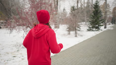 back view of woman jogging outdoors during winter surrounded by snow-covered trees, serene park pathway, evergreen pines, foggy atmosphere, and a residential building in the background