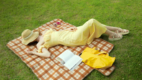 Sexy-woman-in-Yellow-Sundress-Sleeping-Lying-on-Picnic-Plaid-at-Green-Grass-Lawn-at-the-Park-on-Sunny-Day-Covered-Face-with-Straw-Hat