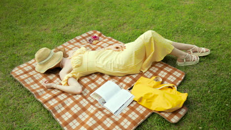 sexy woman in yellow sundress sleeping lying on picnic plaid at green grass lawn at the park on sunny day covered face with straw hat