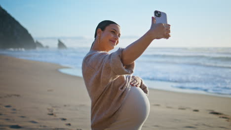 pregnant woman making selfie standing sandy seashore caressing belly close up.