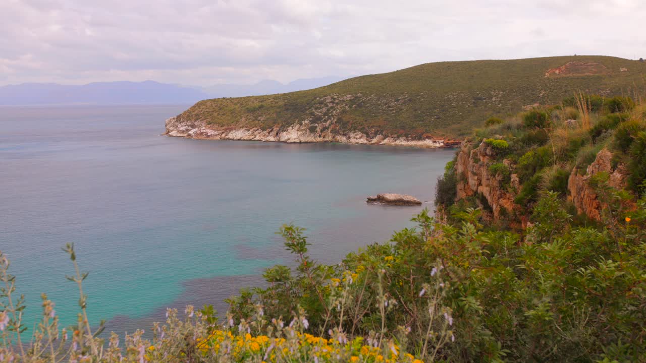 Cliffside View Of Turquoise Bay With Wildflowers In Foreground On Coast Of Sicily In Trapani, Italy. wide shot