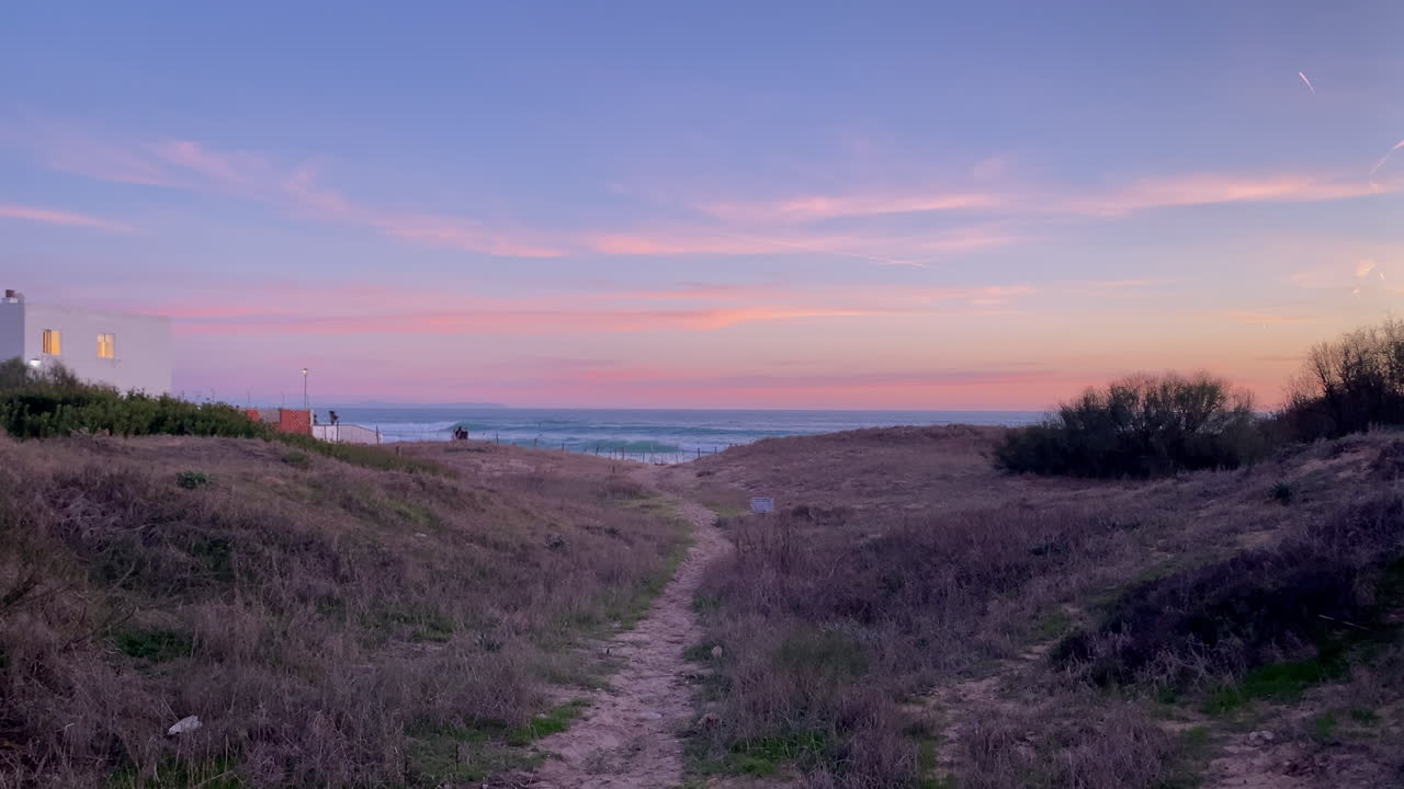 Sandig stig som slingrar sig genom kustdyner mot havet med vågor under en färgstark rosa och blå solnedgångshimmel.