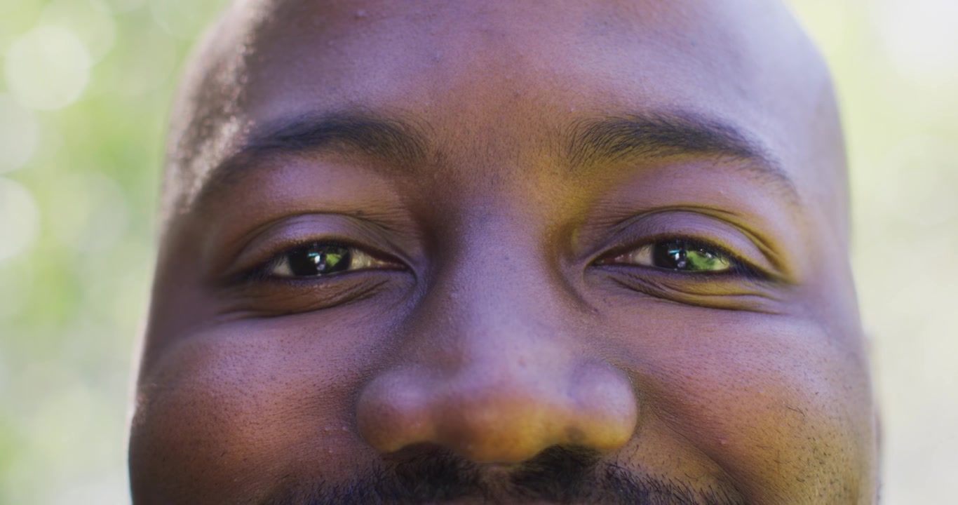 Homem africano adulto interage com a câmera, sorrindo enquanto a câmera se afasta, mostrando o reflexo do olho no parque.