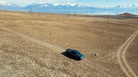 A-truck-driving-along-a-dirt-road-in-an-arid-landscape-with-a-lake-and-snow-mountains-in-the-background---aerial-view