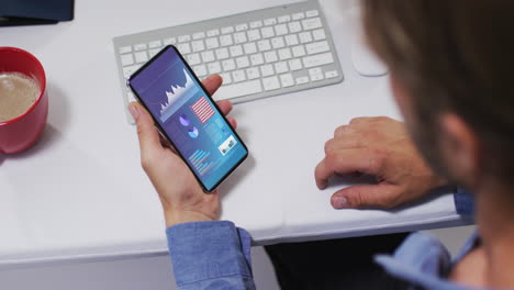 hombre caucásico sentado en un escritorio usando un teléfono inteligente con estadísticas en la pantalla