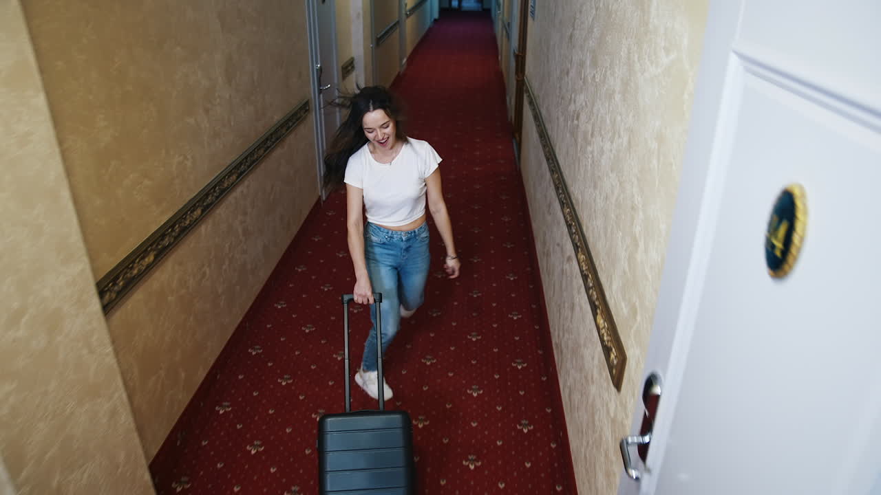 Happy woman with suitcase. Joyful young female traveler walking along the hotel corridor with a luggage and jumping merrily. Top view.