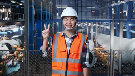 ingeniero asiático con casco de seguridad de pie en una fábrica de fabricación de turbinas eólicas. sonriendo y mostrando un gesto de paz mientras el brazo robótico trabaja