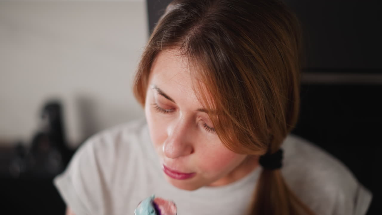 Close up of young woman focusing on colorful ice cream cone with green waffle cone as she licks it while standing in modern kitchen in front of fridge, with kitchen tap slightly visible behind her