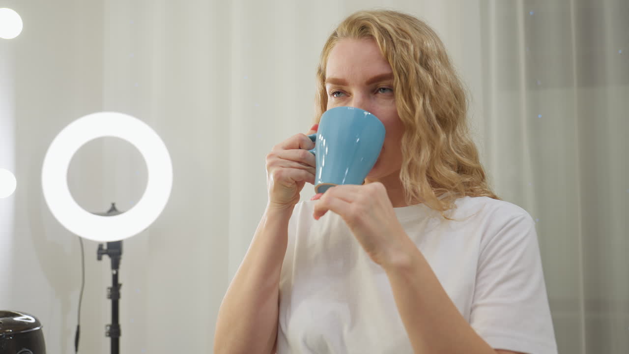 Blond woman wearing white shirt enjoying warm tea from blue cup while standing in bright interior with ring light, mirror, and beauty equipment in background, expressing relaxation and contentment
