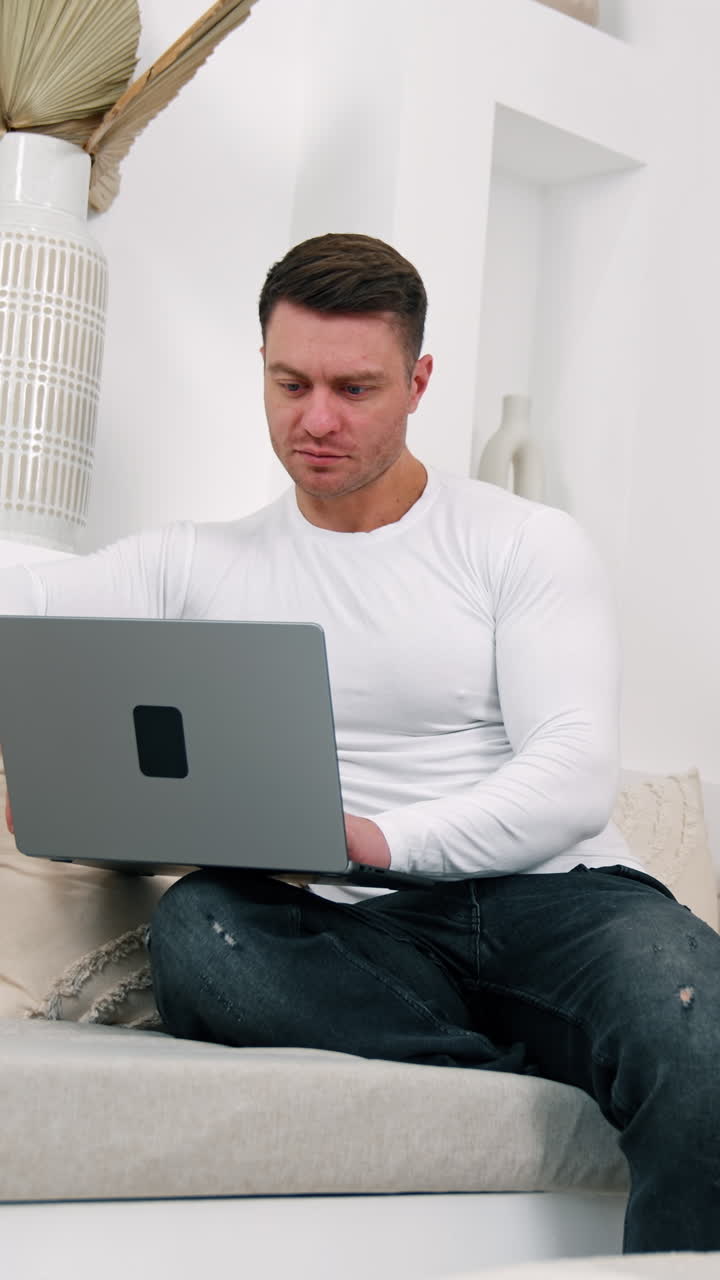 Smiling Caucasian male sits on sofa holding a laptop. Man finishes work, closes his laptop and looks at camera. Vertical video